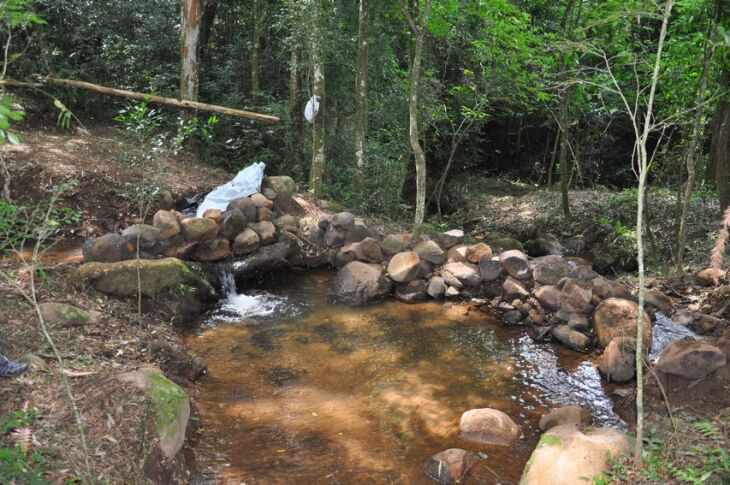 Imagem mostra a remoção de vegetação de sub-bosque, o barramento de curso hídrico com pedras e início da construção de uma área de lazer